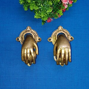 Peut inclure: Deux heurtoirs de porte dorés en forme de mains tenant une sphère. Les heurtoirs sont montés sur un fond bleu.
