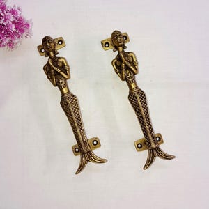 May include: Two bronze mermaid-shaped door handles. The handles feature a mermaid figure with a detailed fishtail and are mounted on rectangular plates with screw holes. The handles are a warm, antique gold color.
