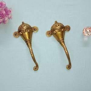 Può includere: Due maniglie per porte dorate a forma di testa di elefante con dettagli intricati. Le maniglie hanno un design curvo e sono montate su una superficie blu chiaro.