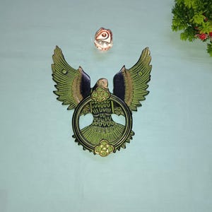 Könnte beinhalten: Ein grüner und goldener Türklopfer aus Metall in Form eines Vogels mit ausgebreiteten Flügeln. Der Vogel hält einen runden Ring in seinen Krallen.