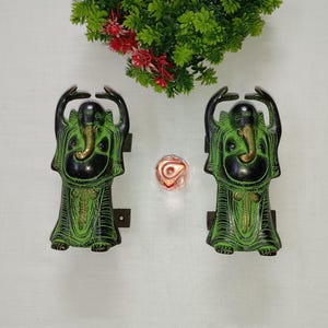 Może przedstawiać: Dwa zielone i złote metalowe uchwyty do drzwi w kształcie hinduskiego bóstwa Ganesha. Uchwyty są uniesione na metalowej podstawie.