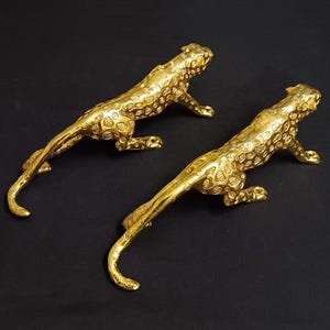 以下が含まれることがあります： 2つの金色の装飾的なチーターの置物。チーターは這う姿勢で、体が長く、尾が曲がっています。チーターは斑点のあるテクスチャ加工された表面で、黒い背景に配置されています。