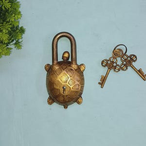 Può includere: Un lucchetto decorativo in ottone a forma di tartaruga, con due chiavi ornate. Il lucchetto ha una superficie strutturata e una serratura. Le chiavi sono collegate da un anello. Gli oggetti sono su una superficie azzurra.