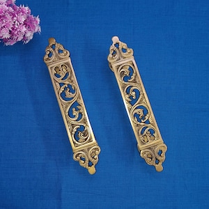 Peut inclure: Deux poignées de porte en laiton ornées de motifs floraux complexes. Les poignées sont de couleur or clair et ont une forme rectangulaire.