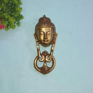 Op de afbeelding: Een gouden messing deurklopper in de vorm van het gezicht van Boeddha. De deurklopper heeft een ring om aan te trekken en een gedetailleerd ontwerp.