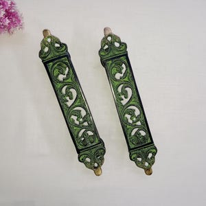 Może przedstawiać: Dwa ozdobne wskaźniki Tory z mosiądzu z zielonymi kwiatowymi wzorami z emalii. Wskaźniki mają około 25 cm długości i ozdobny wierzchołek.