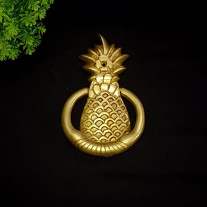 Puede incluir: Un llamador de puerta de piña dorado con un asa de anillo. La piña está detallada con una superficie texturizada y una corona en la parte superior.