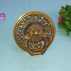 May include: A gold-toned metal door knocker with an intricate design. The knocker is round and has a detailed floral pattern. The knocker is attached to a door with a ring.