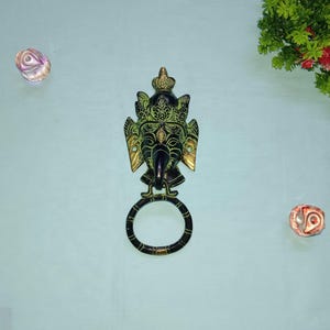 Puede incluir: Un aldaba de latón con la forma de la deidad hindú Ganesha, con una cara detallada, corona y orejas grandes. La aldaba tiene un asa circular y está montada en una pared azul claro. Elementos decorativos se colocan alrededor de la aldaba.