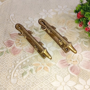 Peut inclure: Deux poignées de porte antiques en laiton avec un motif de crocodile. Les poignées sont ornées d'écailles et d'une tête et d'une queue de crocodile. Elles sont posées sur une nappe à motifs floraux.