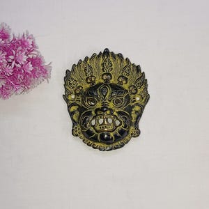 Puede incluir: Máscara decorativa negra y dorada con un diseño detallado y una expresión feroz. La máscara está sobre un fondo blanco, con flores rosas en la esquina superior izquierda.
