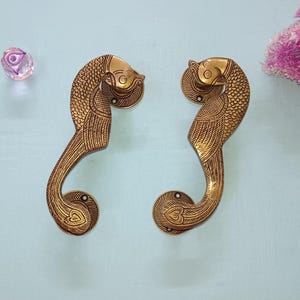 May include: Two ornate, brass-colored door handles in a fish design. The handles are curved with detailed scales and feather patterns. A clear, faceted crystal and pink flowers are in the background.