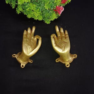 Op de afbeelding: Een paar goudkleurige metalen deurgrepen in de vorm van handen met ingewikkelde ontwerpen. De grepen zijn gemonteerd op een vierkante basis met vier schroeven.