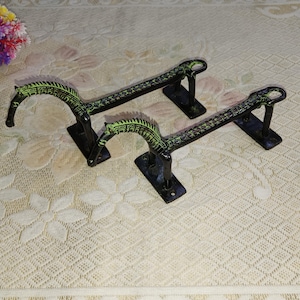 Puede incluir: Dos tiradores de puerta de metal negro con detalles verdes. Los tiradores tienen forma de caballo con la cabeza mirando hacia adelante. Los tiradores están sobre un fondo blanco.