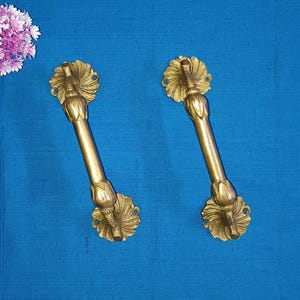May include: Two brass door handles with floral and leaf designs. The handles are gold-colored and have a long, cylindrical shape with decorative ends. The handles are set against a blue background.