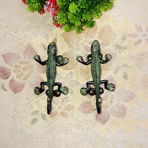 Peut inclure: Deux crochets en métal vert en forme de lézard avec des accents noirs. Les crochets sont montés sur un fond blanc.