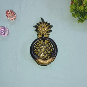 Può includere: Un batacchio per porta a forma di ananas dorato con un anello nero. L'ananas è dettagliato con una superficie testurizzata e un bordo dorato.