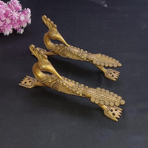 May include: Two antique brass peacock-shaped door handles. The handles are detailed with intricate patterns and designs, featuring a bird's head and a long, decorative body. The handles are golden in color and are placed on a dark surface.