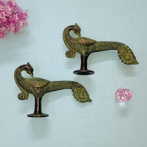 May include: Two antique brass peacock figurines with a green patina. The peacocks are perched on a dark brown base. A pink flower and a small, iridescent glass sphere are also in the image.
