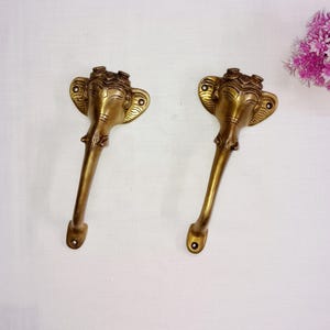 Könnte beinhalten: Zwei goldfarbene Türklinken aus Metall in Form von Elefantenköpfen mit filigranen Details. Die Griffe haben ein langes, geschwungenes Design und sind an einer Wand befestigt.