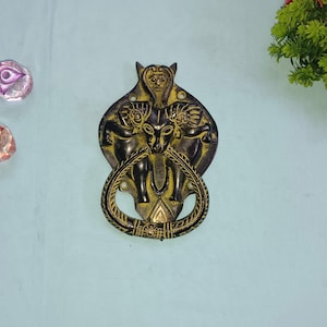 Op de afbeelding: Een gouden deurklopper met een gedetailleerd ontwerp van drie figuren met dierenkoppen. De klopper heeft de vorm van een hart en heeft een touwachtige ring.