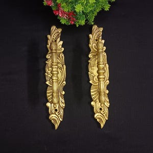 Puede incluir: Dos manijas de puerta doradas ornamentadas con intrincados diseños florales y de volutas. Cada manija presenta un agarre central vertical y adornos decorativos. Las manijas se muestran sobre un fondo oscuro.