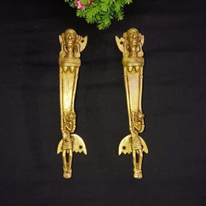 May include: A pair of gold-toned door handles with intricate designs featuring human figures. The handles are made of metal and have a polished finish.