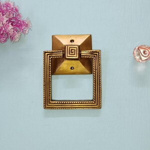 May include: A gold-colored, square door knocker with a geometric design and a spiral center. The knocker is mounted on a light blue surface, with a clear glass knob and pink flowers nearby.