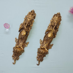 Könnte beinhalten: Zwei verzierte, goldfarbene Wandleuchter mit der hinduistischen Gottheit Ganesha. Jeder Leuchter hat aufwendige Details und eine geschwungene, dekorative Basis. Der Hintergrund ist hellblau mit einer rosa Blume und einem klaren, rosa Kristall.