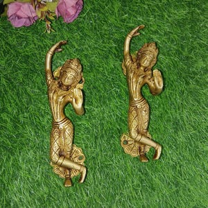 Op de afbeelding: Twee goudkleurige metalen deurgrepen in de vorm van dansende figuren. De figuren staan in een sierlijke houding met hun armen boven hun hoofd. De grepen hebben ingewikkelde details en een decoratieve basis.