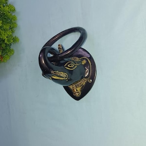 Puede incluir: Anillo para toalla de metal negro en forma de cabeza de elefante con un acento dorado. El anillo es circular y la cabeza de elefante está detallada con tallas intrincadas.