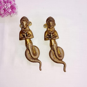 May include: Two ornate brass door handles shaped like a woman with a snake tail. The woman is in a prayer position with her hands clasped together. The handles are a gold colour.