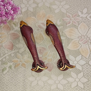 Pode incluir: Duas maçanetas de porta de latão ornamentadas em forma de peixe com acabamento vermelho. As maçanetas têm uma moldura dourada e são montadas em um fundo branco.