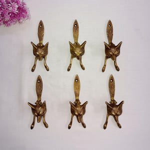 Può includere: Sei ganci dorati a forma di testa di volpe con una lunga coda. I ganci sono montati su una parete bianca.