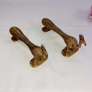 Pode incluir: Duas maçanetas de porta de latão ornamentadas em forma de pavão com a cabeça virada para o lado. As maçanetas são detalhadas com entalhes intrincados e têm um acabamento dourado.