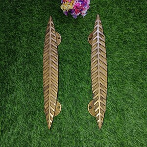 Może przedstawiać: Dwa złote uchwyty do drzwi w kształcie liścia z geometrycznym wzorem. Uchwyty znajdują się na zielonym sztucznym tle trawy.