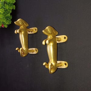 Può includere: Due maniglie per porte a forma di cane bassotto dorate. Le maniglie sono montate su uno sfondo bianco.