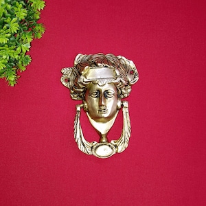 Może przedstawiać: Złoty klamka do drzwi z detalicznym wzorem twarzy kobiety. Klamka ma ozdobny pierścień wokół głowy kobiety i pierścień wokół szyi.