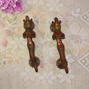 May include: A pair of ornate brass door handles shaped like a woman with a snake tail. The handles are a reddish brown colour with gold accents.