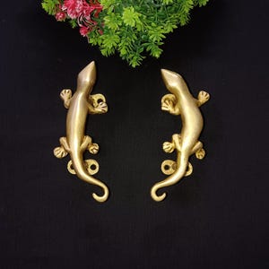 Peut inclure: Une paire de poignées de porte en laiton doré en forme de lézards. Les poignées sont détaillées avec des écailles texturées et ont une finition lisse et polie.