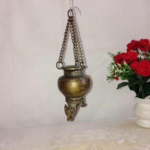 May include: A gold-toned metal incense burner with a round body and three legs. The burner is suspended from a chain and has a decorative design.