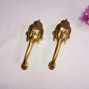 Peut inclure: Deux poignées de porte dorées avec un motif de tête de Bouddha. Les poignées sont longues et courbées, avec un visage et des cheveux détaillés.
