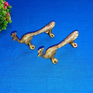 May include: Two brass peacock door handles with intricate details. The handles are in the shape of a peacock with its tail feathers spread out. The handles are a gold colour and have a polished finish.