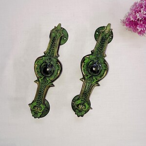 Può includere: Due maniglie per porte ornate in verde e oro con intricati motivi floreali. Le maniglie sono in metallo e hanno un centro nero.