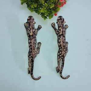 Puede incluir: Un par de ganchos de metal con estampado de leopardo negro y blanco. Los ganchos tienen forma de leopardos trepando por una pared.