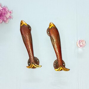 Può includere: Due maniglie per porte a forma di pesce color bronzo. Le maniglie presentano un motivo a scaglie strutturato e una testa e una coda color oro. Le maniglie sono montate su una base rotonda color bronzo.