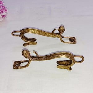 May include: Two gold-toned metal monkey door handles with a decorative design. The handles are curved and have a monkey head at each end.