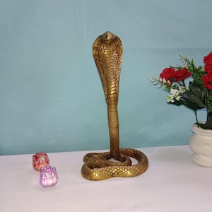 Op de afbeelding: Een gouden messing beeld van een cobra met zijn kap omhoog. Het beeld staat op een wit oppervlak met twee kleine, doorzichtige glazen voorwerpen op de voorgrond.