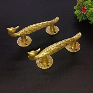 Puede incluir: Dos manijas de puerta doradas y ornamentadas. Cada manija presenta un diseño similar a un pájaro con un agarre curvo en forma de pluma y una base circular. Las manijas están sobre un fondo oscuro.