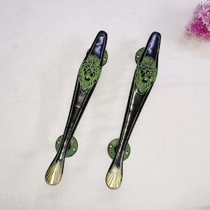 Peut inclure: Deux poignées de porte en métal noir avec des motifs de tête de lion verts. Les poignées ont une extrémité arrondie et pointue et une base circulaire.